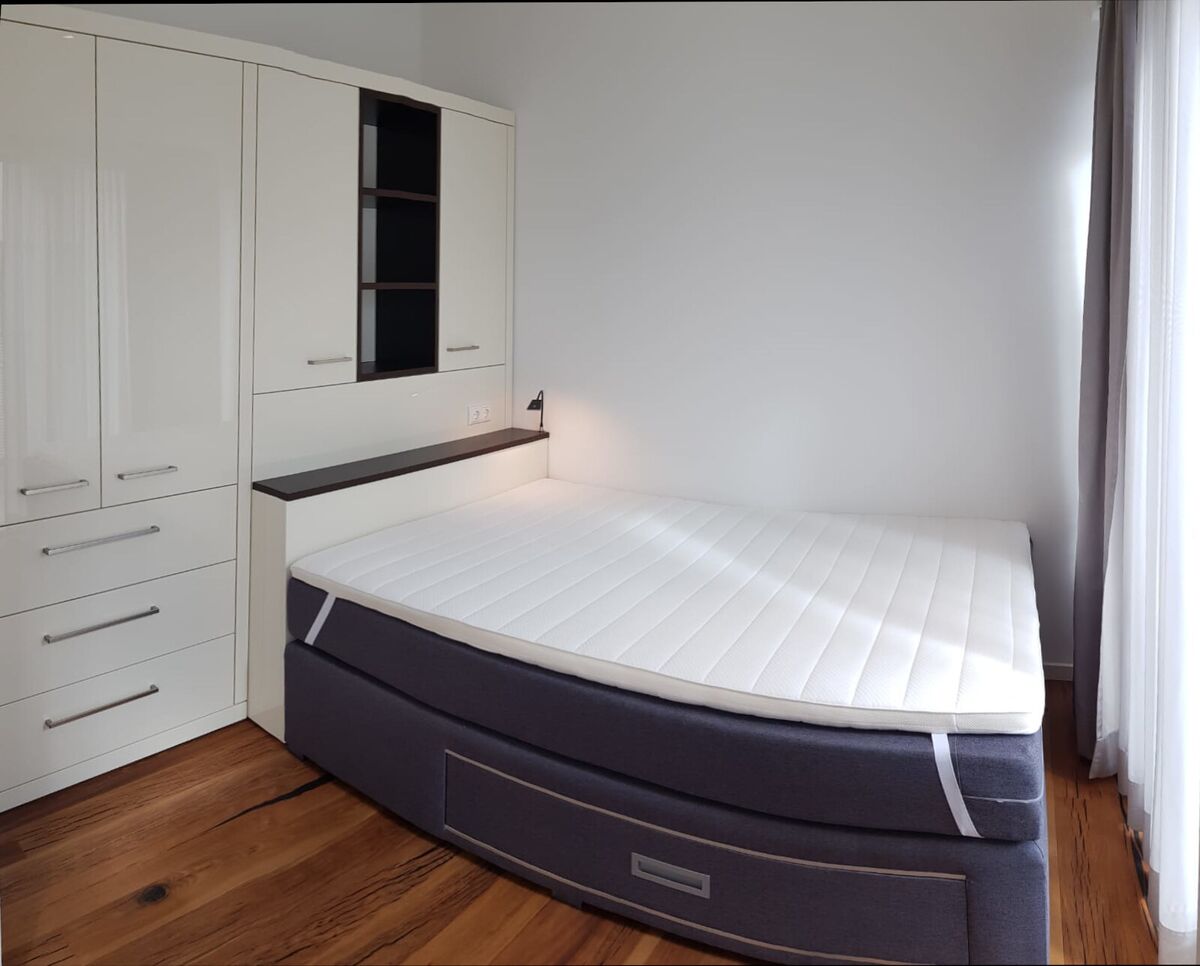 Weißes Bett in der Raumecke mit heller Rückwand und Ablage